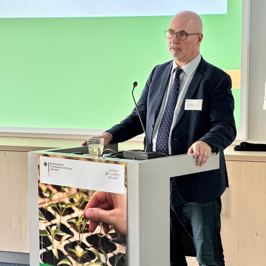 Ein Mann steht im Anzug hinter einem Rednerpult mit Mikrofon und hält ein Grußwort.