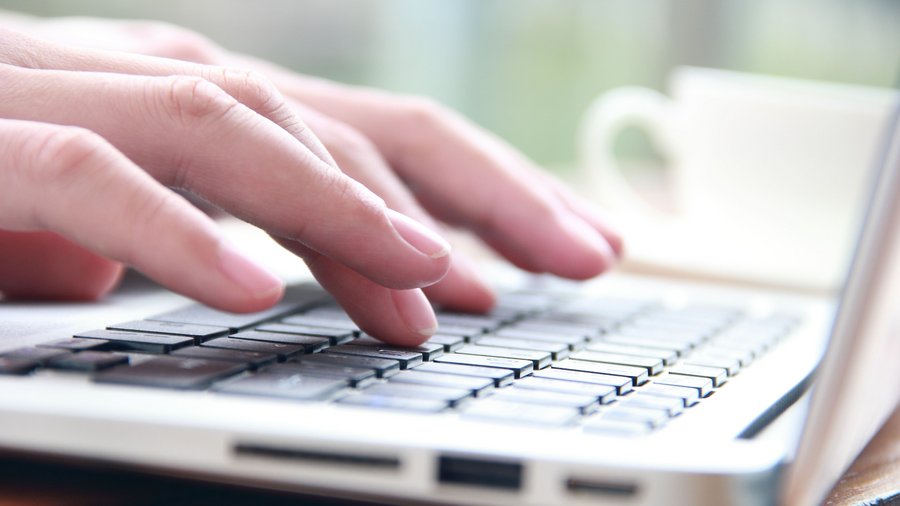 Das Bild zeigt weibliche Hände von der rechten Seite, die auf einer Laptoptastatur tippen. Im Hintergrund sieht man eine weiße Tasse neben dem Laptop stehen..